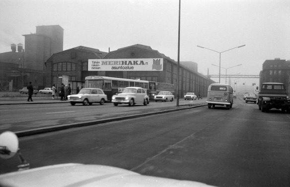 1970 . Näkymä sumuiselta Sörnäisten rantatieltä lounaaseen. Etualalla autoliikennettä, taustalla teollisuusrakennus, jonka päädyssä kyltti, jossa teksti: Tähän rakennetaan MERIHAKAn asuntoalue. Syksy 1970.